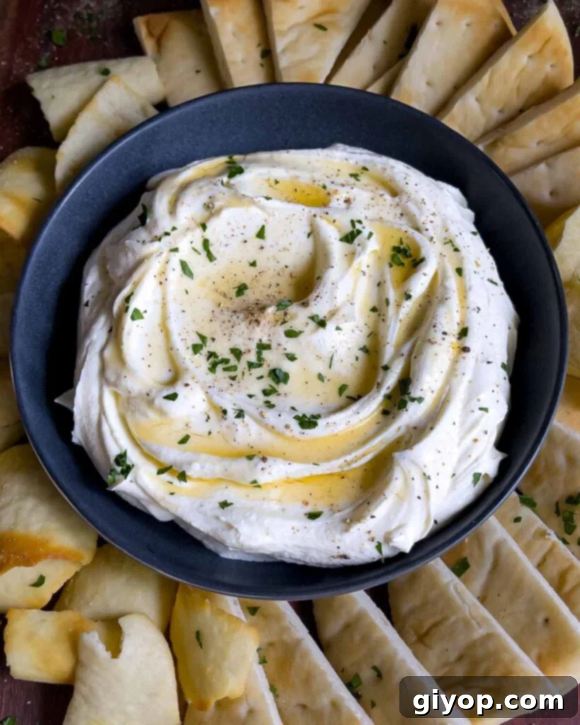 Whipped feta garnished with honey, chopped parsley, and black pepper in a dark bowl surrounded by pita chips and pita bread.