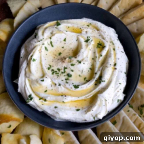 Whipped feta garnished with honey, chopped parsley, and black pepper in a dark bowl surrounded by pita chips and pita bread.