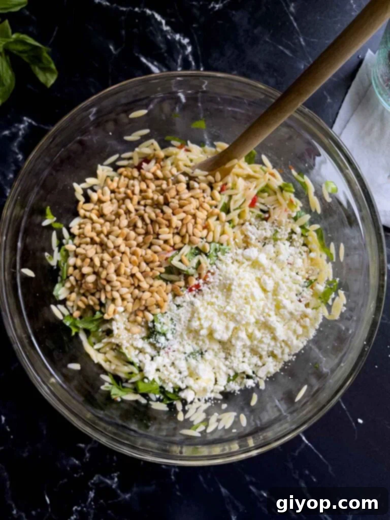 Toasted pine nuts and crumbled feta cheese being added to the spinach and feta orzo salad in a glass bowl on a dark surface, enhancing texture and flavor.