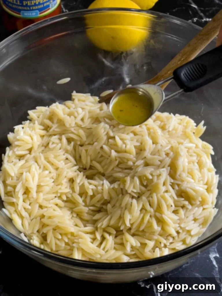 Drained orzo pasta being tossed with a small amount of olive oil in a clear glass bowl, allowing it to cool without sticking.