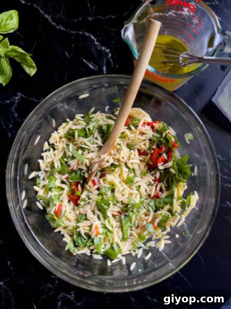 Chopped vegetables and herbs being added to the cooled orzo pasta in a glass bowl, with a measuring cup of orzo salad dressing nearby.