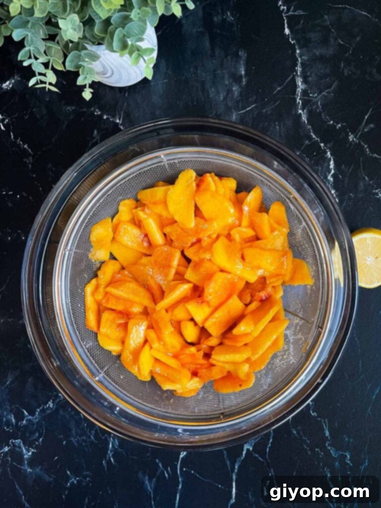 Freshly sliced and chunked peaches, glistening with lemon juice, draining in a fine mesh strainer over a glass bowl, ready for pie filling.