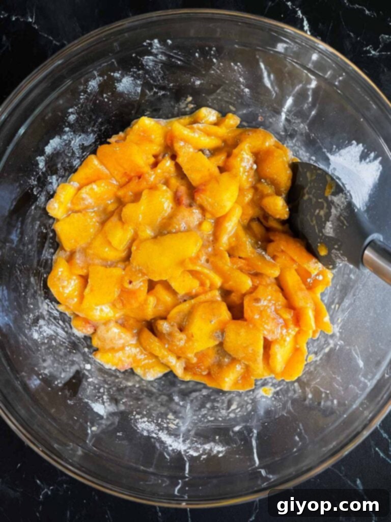 A vibrant display of fresh peach slices and chunks, evenly coated with a sweet and spiced dry ingredient mixture, in a clear glass bowl.