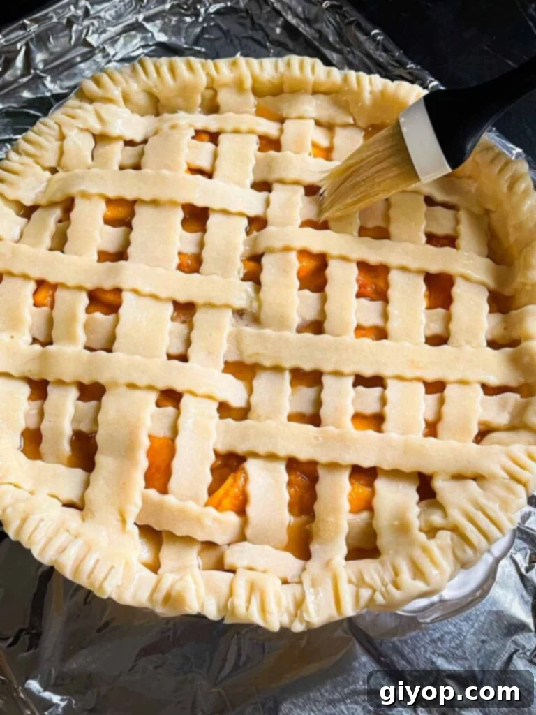 A chef's hand delicately brushing an egg wash over the intricate lattice crust of an unbaked peach pie, preparing it for a golden finish.