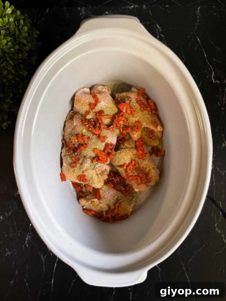 Raw chicken thighs and chopped sun-dried tomatoes neatly arranged in the bottom of a slow cooker before cooking.