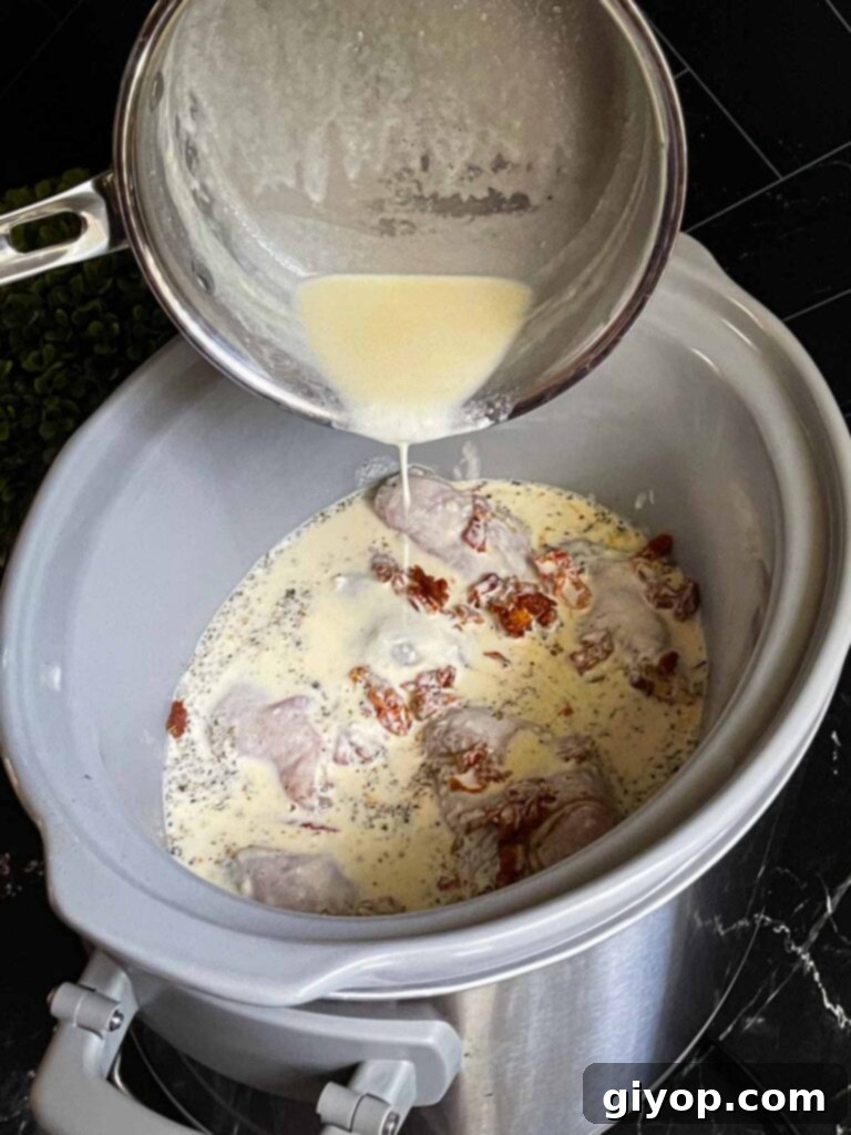 The creamy Tuscan sauce being poured over the raw chicken and sun-dried tomatoes already in the slow cooker.
