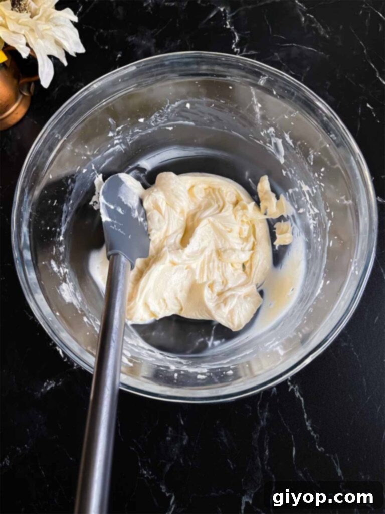 A block of softened cream cheese, beaten smooth and creamy, resting in a glass mixing bowl on a dark surface, awaiting further ingredients.