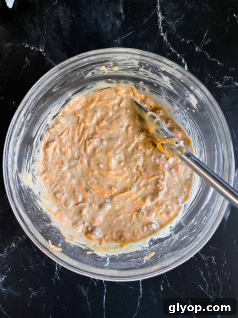 The fully mixed sweet potato cake batter, speckled with orange sweet potato shreds and pecan pieces, sitting in a large glass mixing bowl on a dark surface.