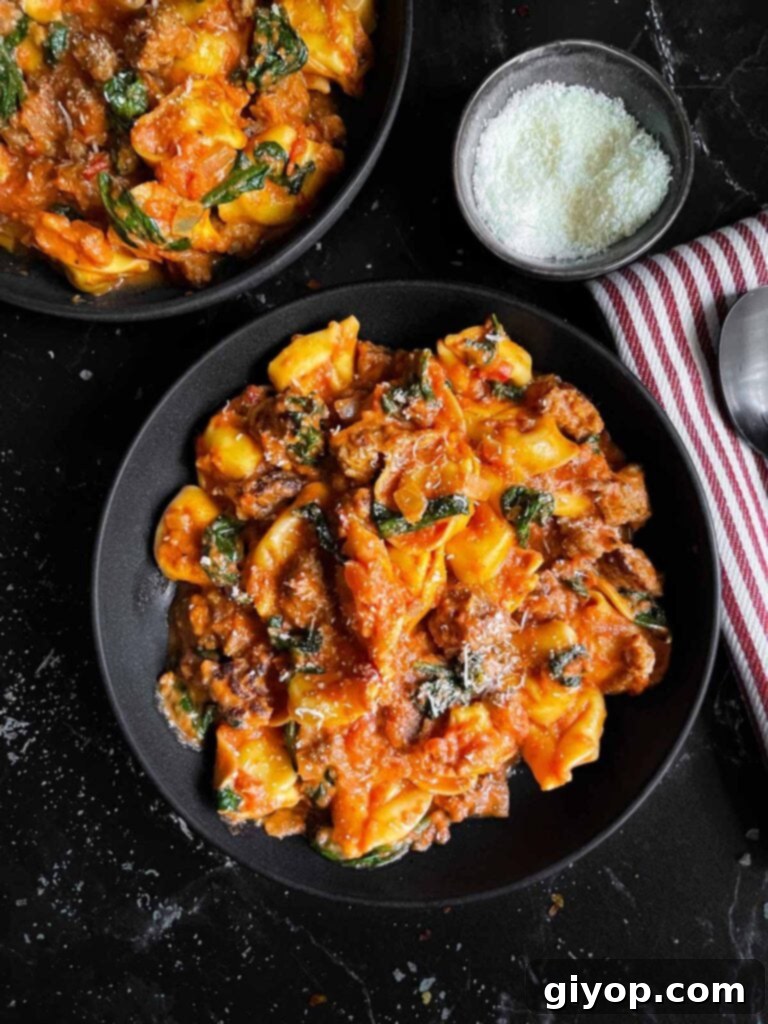 Creamy cheese tortellini with Italian sausage and wilted spinach in a dark ceramic bowl, placed on a contrasting dark surface, ready to be served.