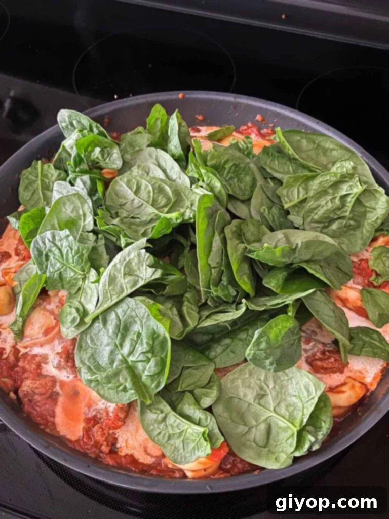 Fresh spinach being added to the one-pot tortellini and Italian sausage dish in a skillet.