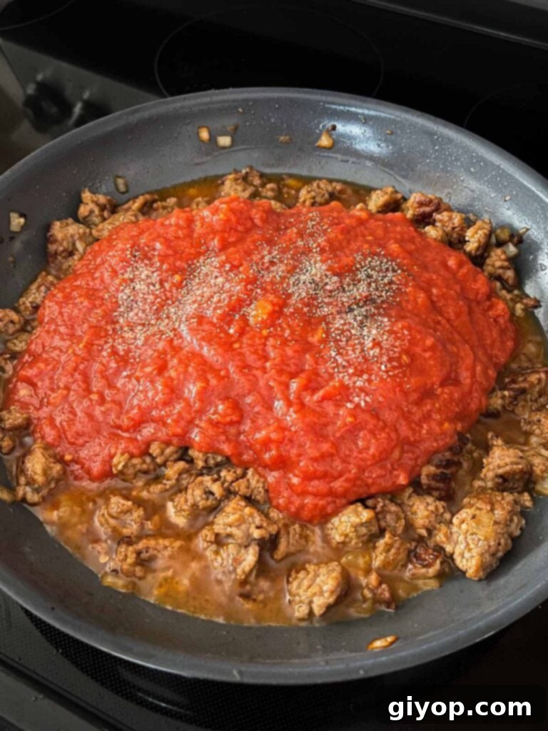 Crushed tomatoes being poured into the skillet with Italian sausage and aromatics, forming the base of the sauce.