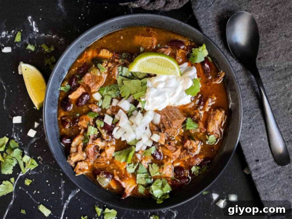 Smoked pork chili garnished with cilantro, onion, lime, sour cream, and onion in a dark bowl.