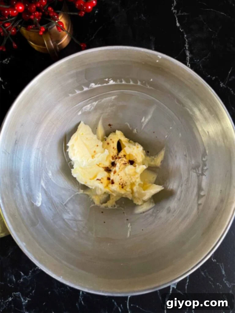 Vanilla extract being added to the creamy, whipped butter and powdered sugar mixture in a shiny metal mixing bowl, ready for the next step.