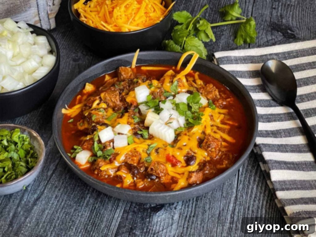 Brisket chili garnished with onions, cilantro, and cheddar cheese in a bowl sitting on a wooden background.