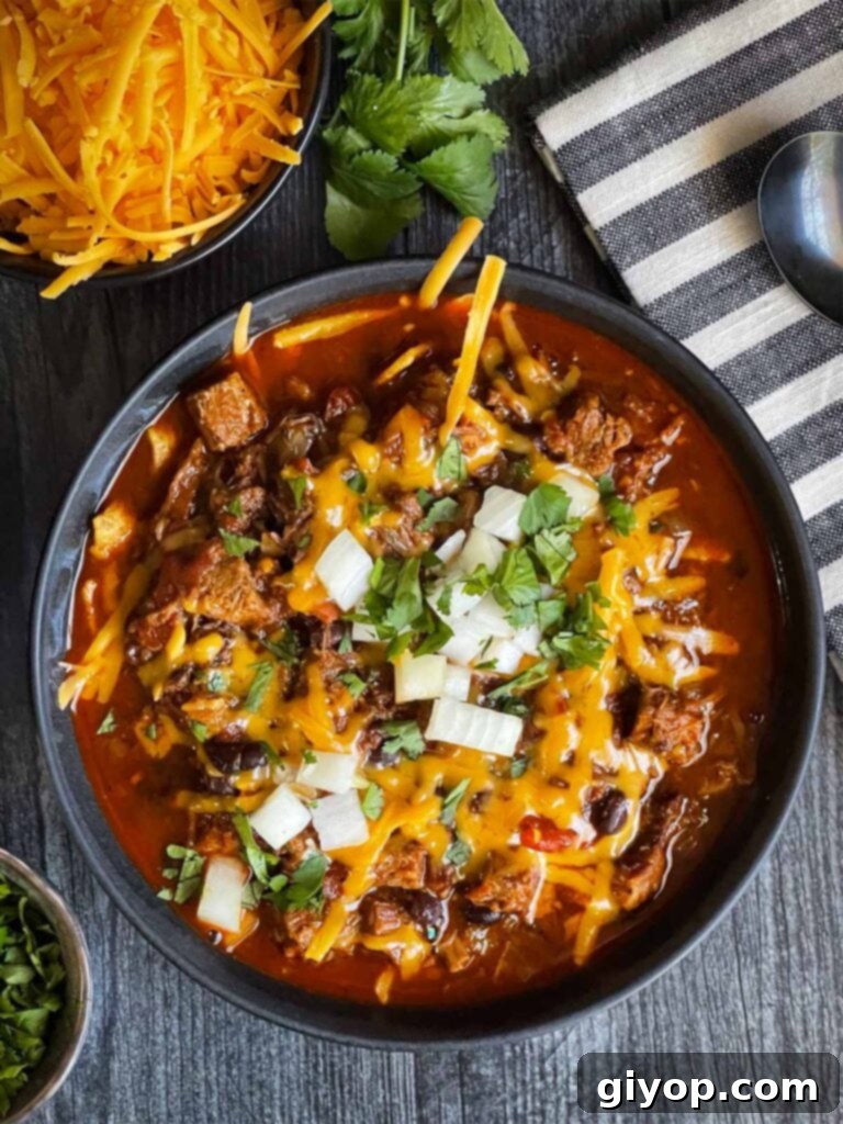 Brisket chili garnished with onions, cilantro, and cheddar cheese in a bowl sitting on a wooden background.