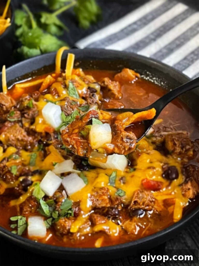 Brisket chili garnished with onions, cilantro, and cheddar cheese in a bowl sitting on a wooden background.