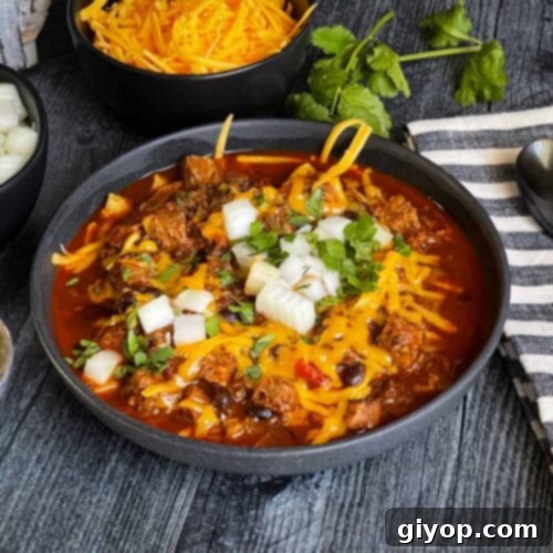 Brisket chili garnished with onions, cilantro, and cheddar cheese in a bowl sitting on a wooden background.