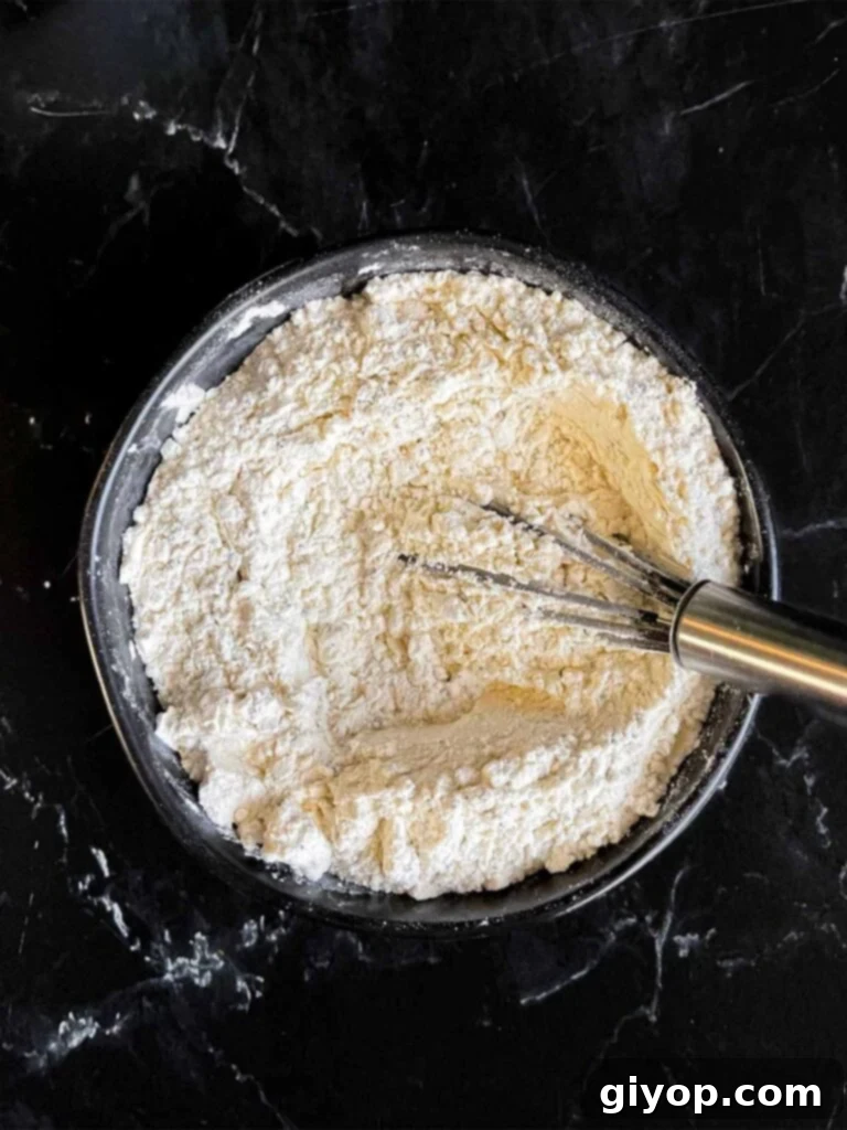 Dry ingredients for lemon poppy seed muffin recipe, including flour, baking powder, kosher salt, and poppy seeds, whisked together in a dark bowl on a dark surface, ready for the next step.