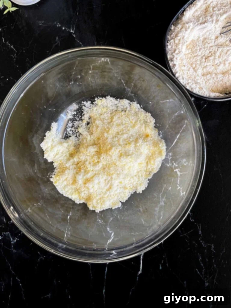 Granulated sugar and vibrant lemon zest are rubbed together in a clear glass mixing bowl, placed on a dark surface, illustrating the process of infusing the sugar with citrus aroma.