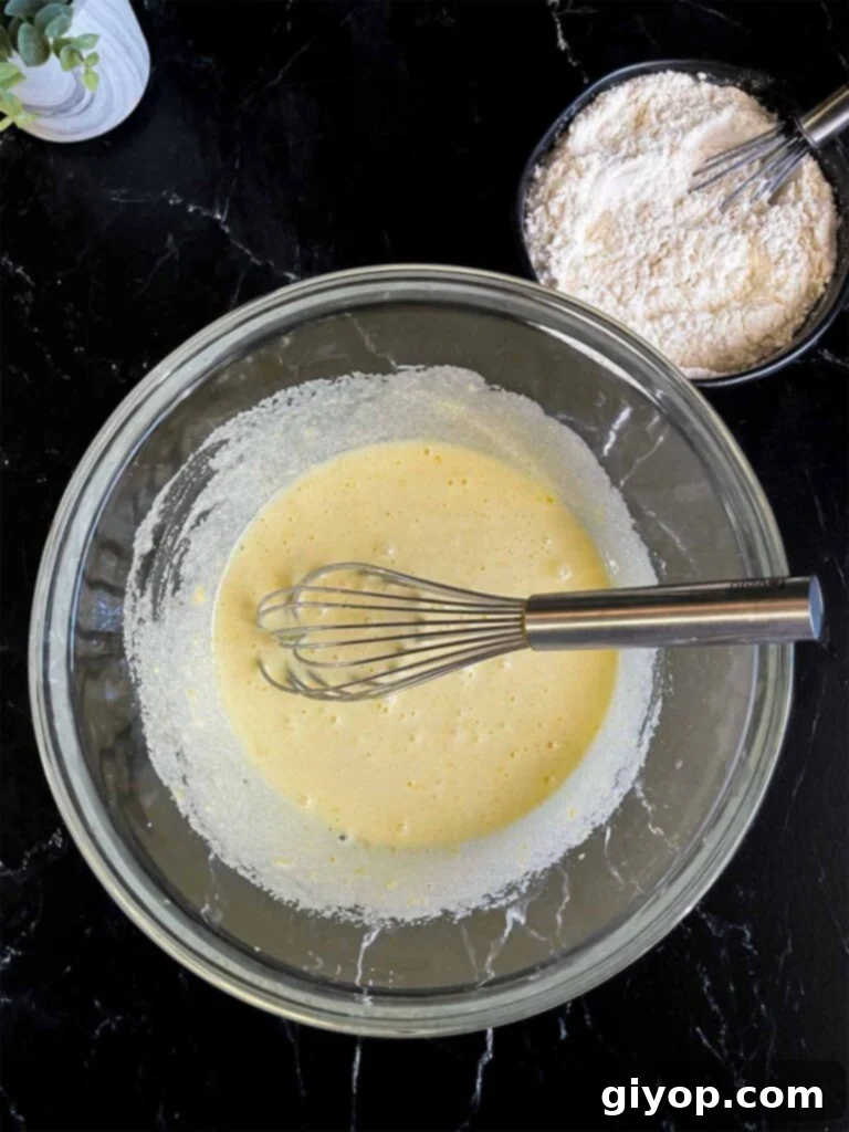Eggs being whisked into granulated sugar infused with lemon zest in a clear glass mixing bowl on a dark surface, showing the emulsification process.