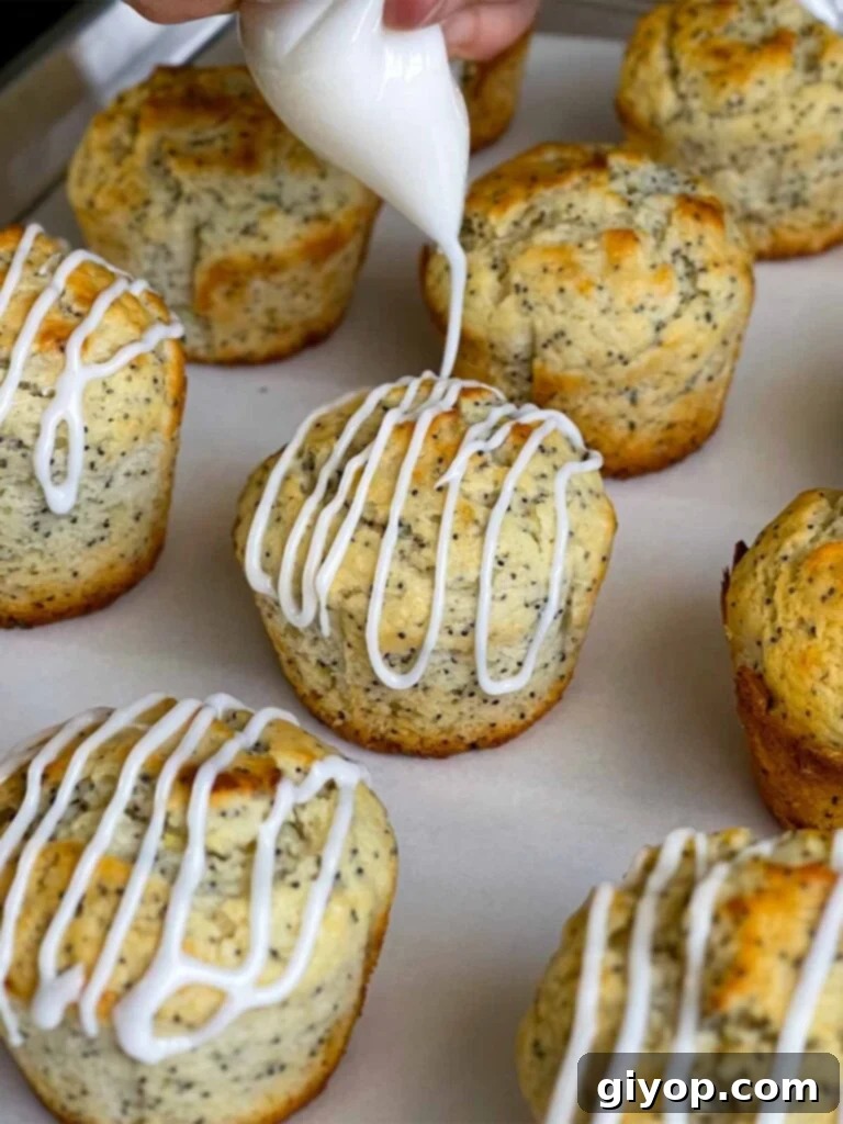A smooth lemon glaze is being drizzled artfully over warm lemon poppy seed muffins on a parchment paper-lined baking sheet, adding a sweet and tangy finish.