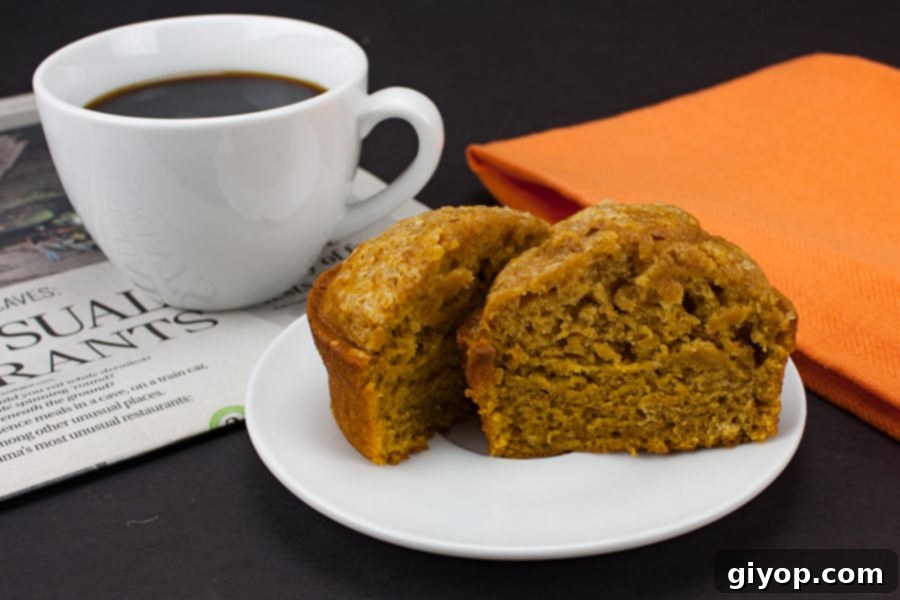 A freshly baked bakery-style pumpkin muffin, sliced in half, rests on a white plate next to a steaming white coffee mug, highlighting its perfect texture and a cozy autumn vibe.
