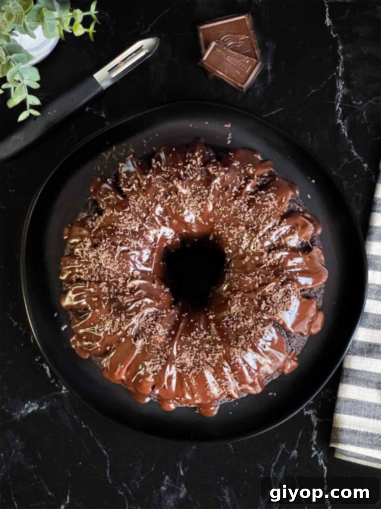 A finished chocolate bundt cake generously topped with shiny chocolate ganache and elegantly garnished with chocolate shavings, presented on a dark plate.