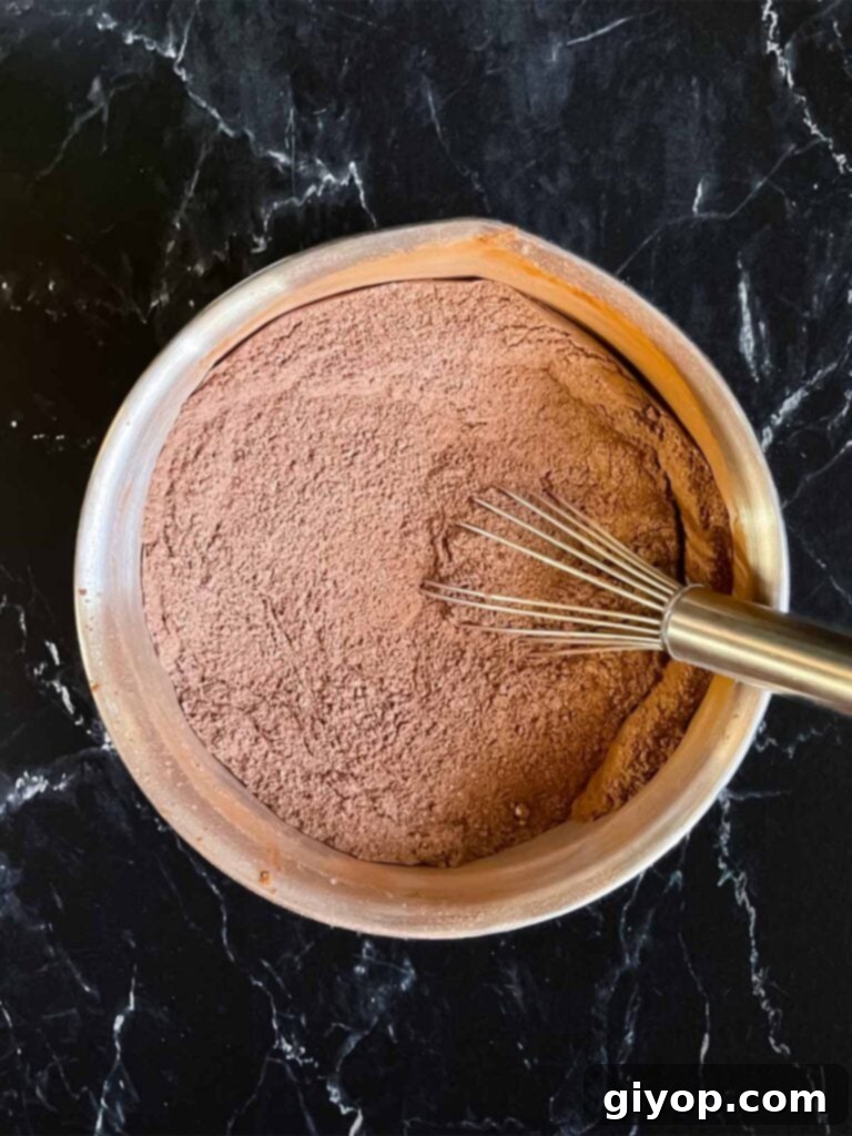 Dry ingredients for chocolate bundt cake whisked together in a metal mixing bowl on a dark surface.