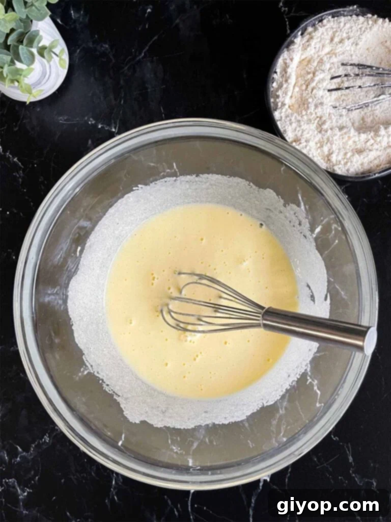 Granulated sugar and eggs being whisked together in a glass mixing bowl until thick and pale, with the dry ingredients in a separate dark bowl on a dark surface.