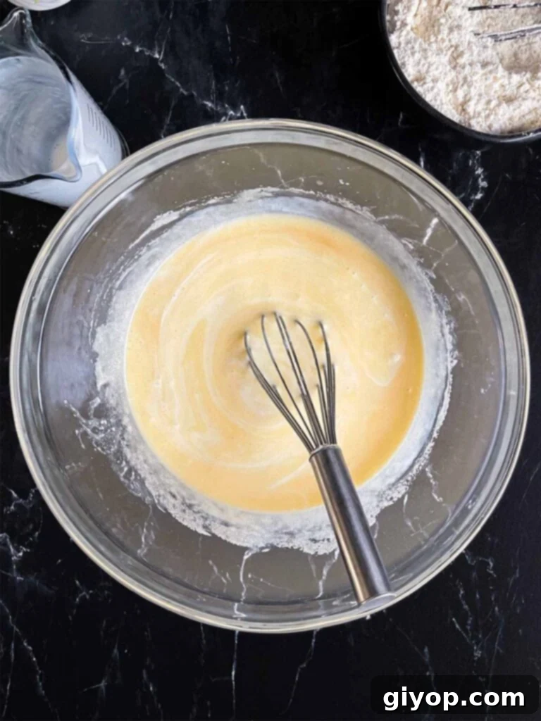 The combined wet ingredients for raspberry muffins, including eggs, sugar, oils, buttermilk, and vanilla, in a glass mixing bowl, with dry ingredients waiting in a separate dark bowl.