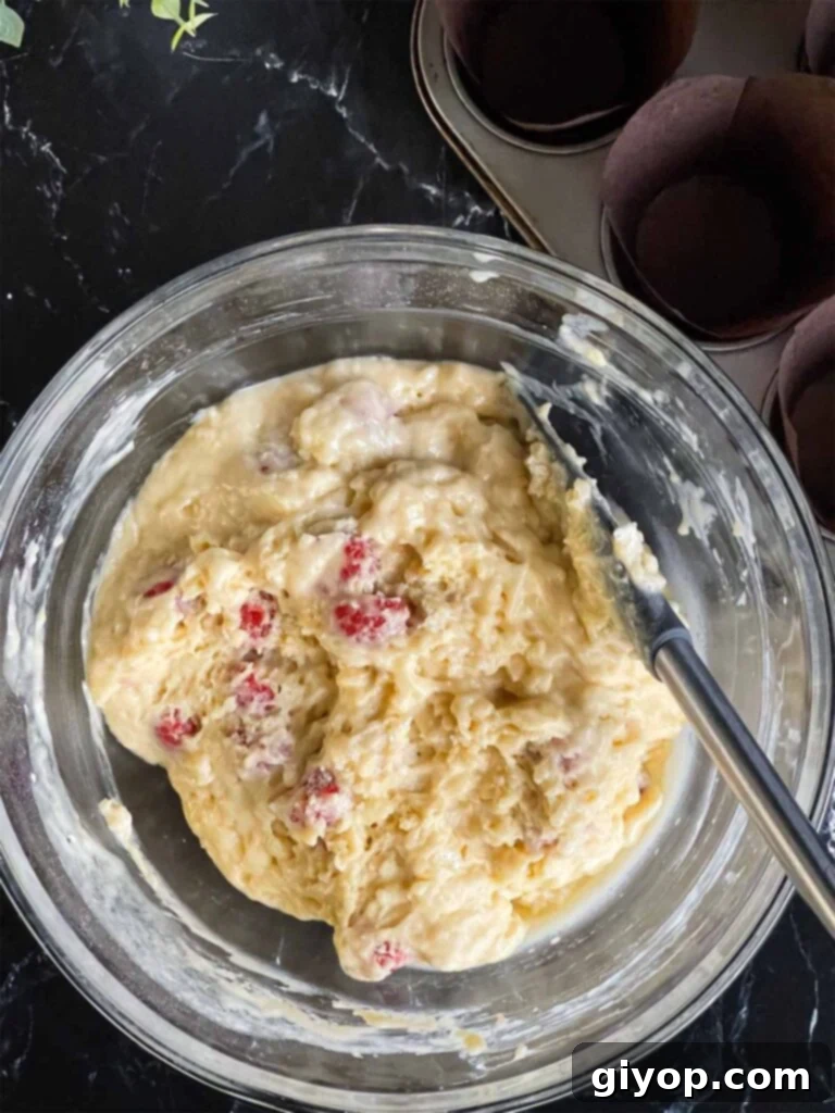The raspberry muffin batter, displaying a slightly lumpy texture and even distribution of raspberries, rests in a glass mixing bowl on a dark surface after 15 minutes.
