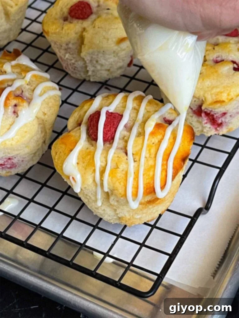 A steady stream of bright lemon glaze is drizzled from a piping bag over golden-brown raspberry muffins cooling on a wire rack, set over a parchment-lined baking sheet.