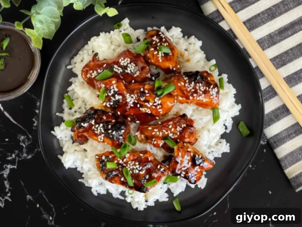Bourbon chicken on a bed of white rice on a dark plate, garnished with sesame seeds and green onion.