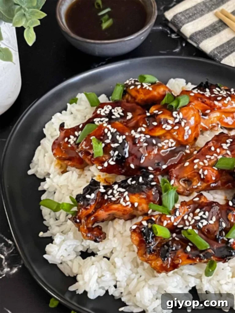 Bourbon chicken on a bed of white rice on a dark plate, garnished with sesame seeds and green onion.