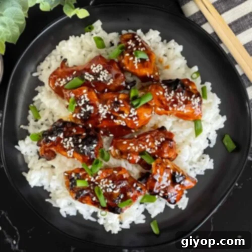 Bourbon chicken on a bed of white rice on a dark plate, garnished with sesame seeds and green onion.