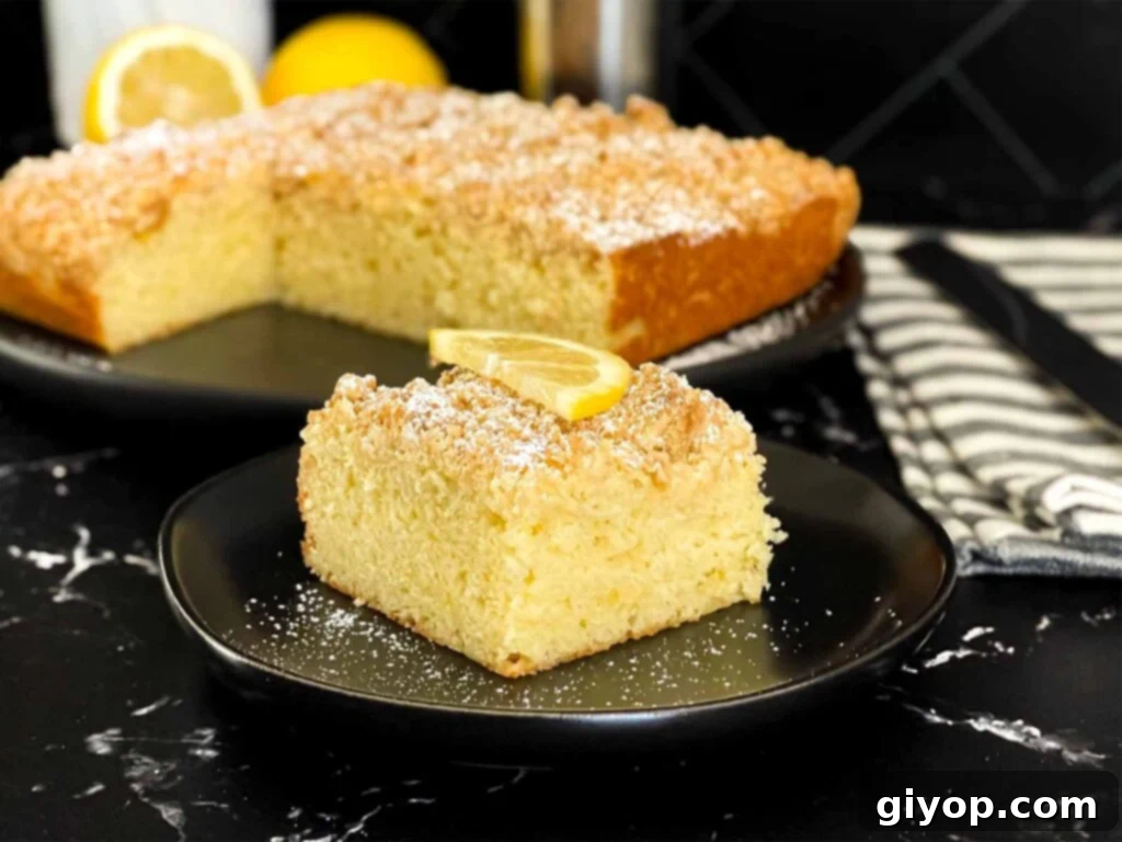 A slice of lemon crumb cake dusted with powdered sugar and garnished with a lemon slice on a dark plate on a dark surface.