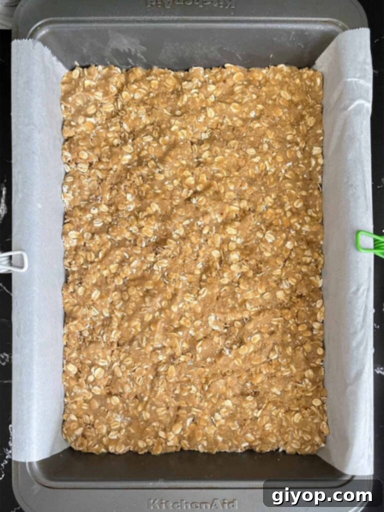 Chewy Oatmeal Fudge Bars 6 A generous layer of oatmeal cookie dough perfectly pressed into the base of a 9x13 baking pan, ready for the fudge filling, with parchment paper lining for easy removal.