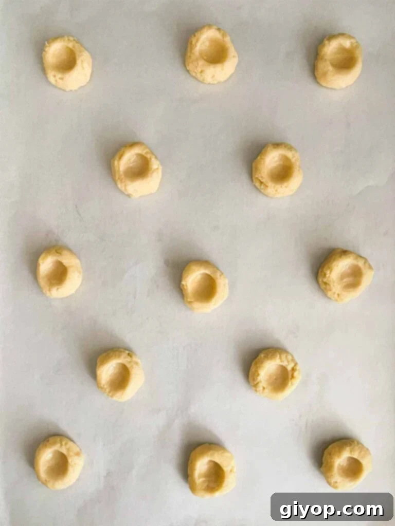 Lemon thumbprint cookie dough balls, each with a neat indentation, resting on parchment paper on a baking sheet, ready for baking.