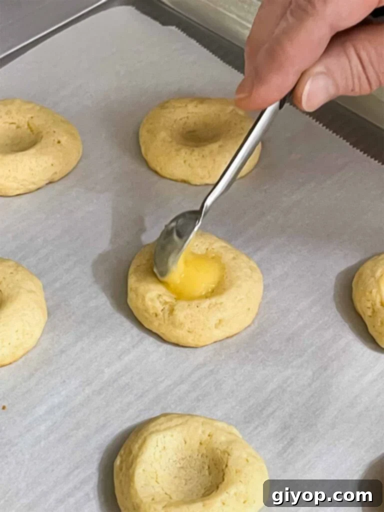 Smooth lemon curd is carefully spooned into the thumbprint indentations of partially baked cookies on a parchment-lined baking sheet.