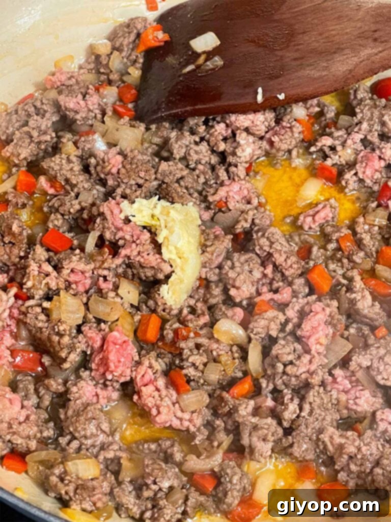 Minced garlic being added to the browning ground beef, red bell peppers, and onion mixture in a skillet.