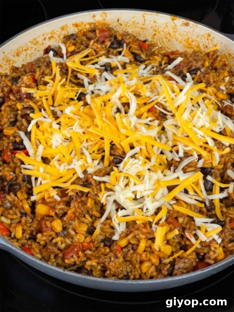 Adding shredded cheese to the hot Mexican ground beef and rice mixture in a skillet, ready to melt.