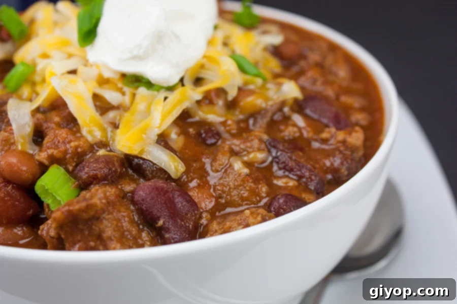 A vibrant bowl of thick and hearty homestyle chili, richly garnished with freshly chopped green onions, a generous sprinkle of shredded cheddar cheese, and a dollop of creamy sour cream, ready to be enjoyed.