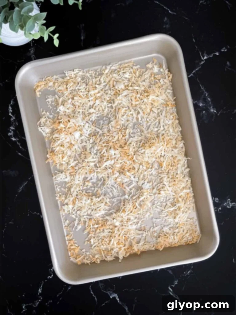 Velvet Coconut Dream 5 Sweetened shredded coconut spread in a single layer on a metal baking sheet, ready for toasting, on a dark surface.