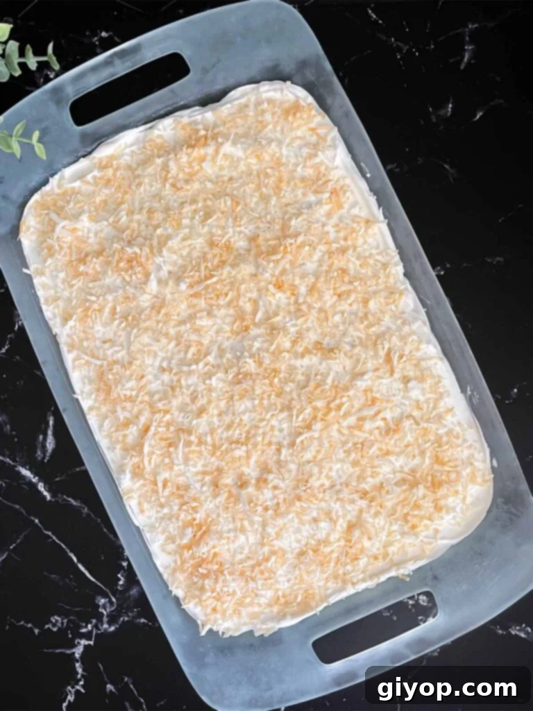 Velvet Coconut Dream 10 The final whipped topping layer spread perfectly over the coconut cream lush, garnished with toasted shredded coconut, in a glass baking pan on a dark surface.