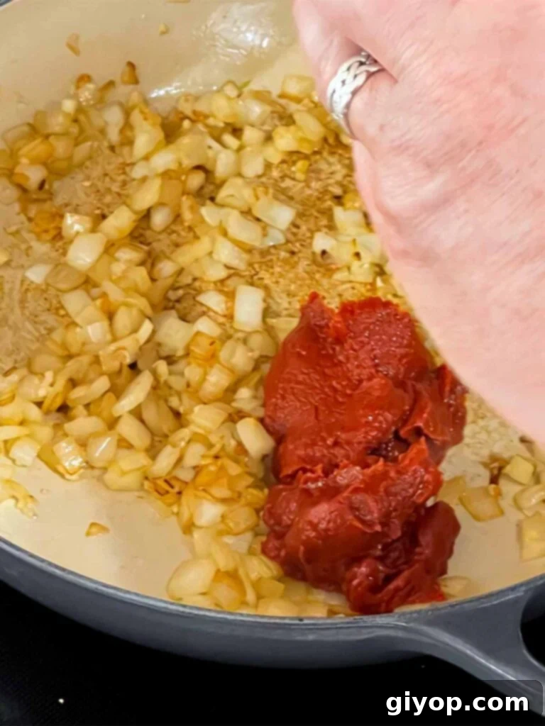 Tomato paste being added to a skillet with cooked onions and garlic.