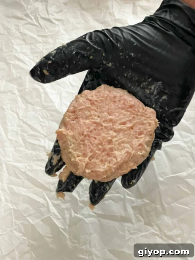 A raw chicken burger patty held gently in a black gloved hand.