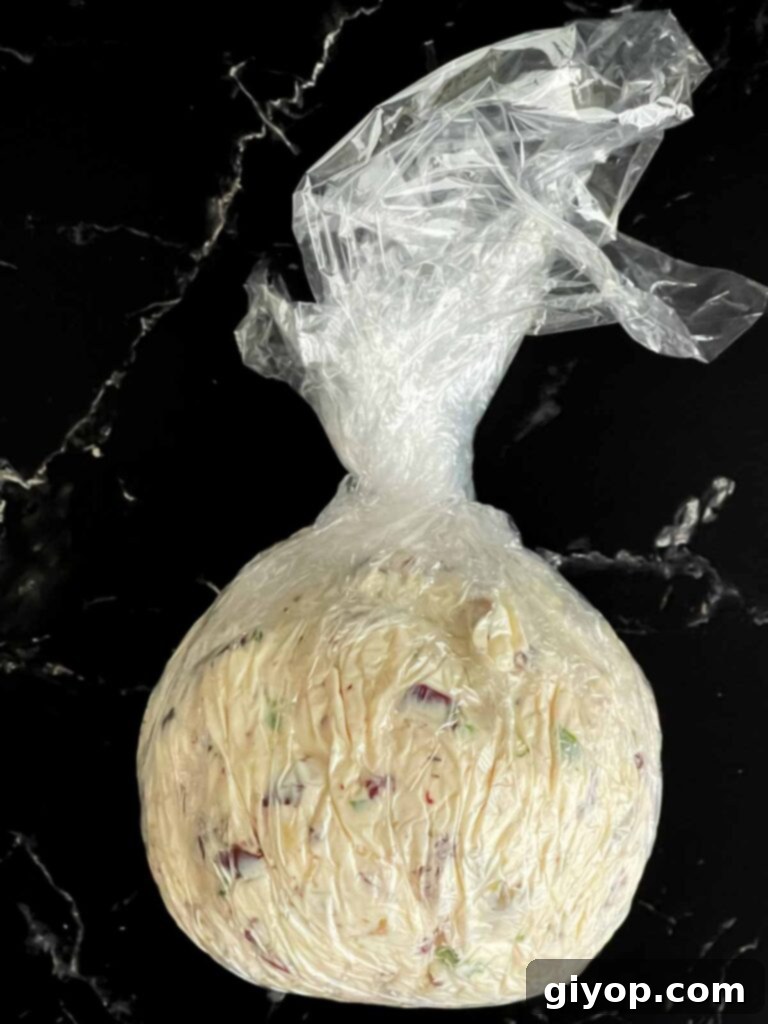 Mediterranean Olive Cheese Ball 7 The olive cheese ball perfectly formed into a round shape and tightly encased in plastic wrap, resting on a dark surface.