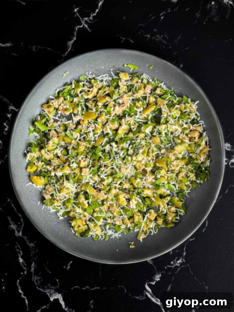 Mediterranean Olive Cheese Ball 8 The vibrant olive cheese ball coating ingredients, including chopped green olives, parsley, and cheese, mixed in a shallow bowl on a dark surface.