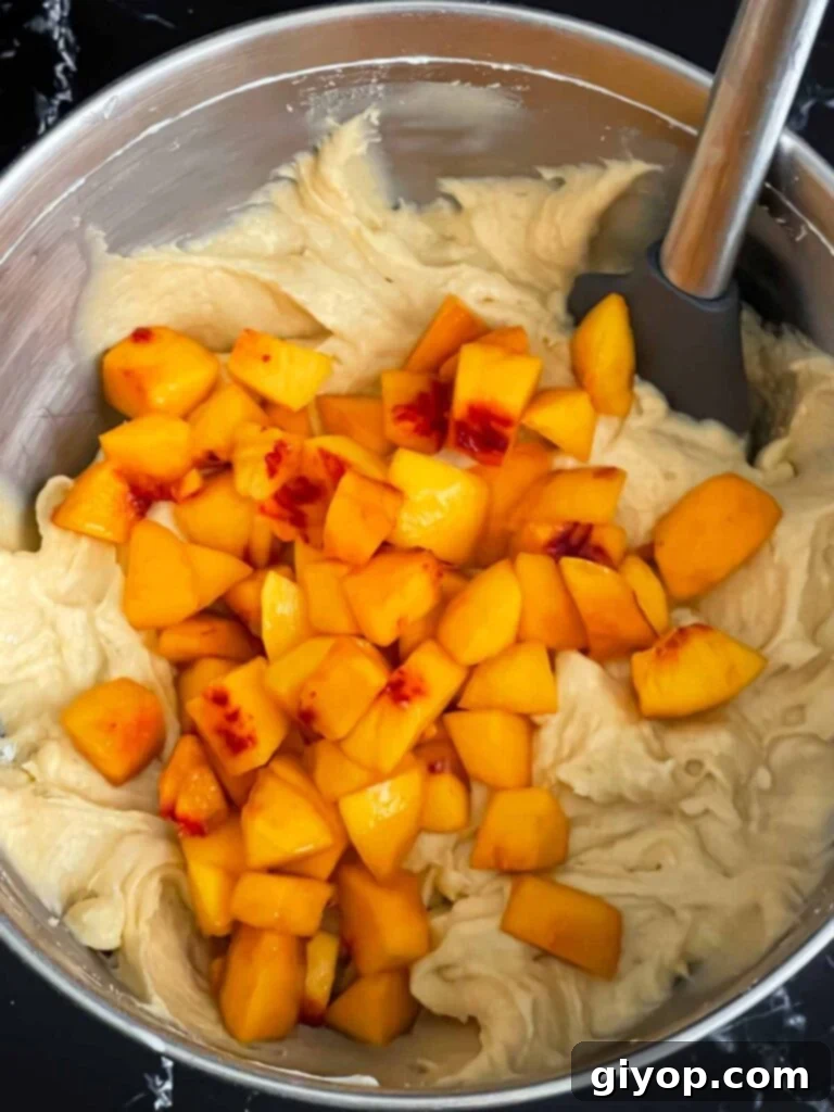 Fresh peach chunks gently folded into the peach pound cake batter in a metal mixing bowl, adding bursts of fruity flavor.