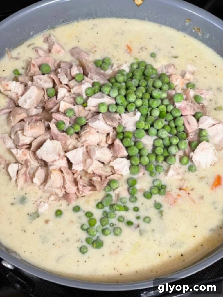 Diced chicken and frozen peas being added to the creamy sauce in a skillet.